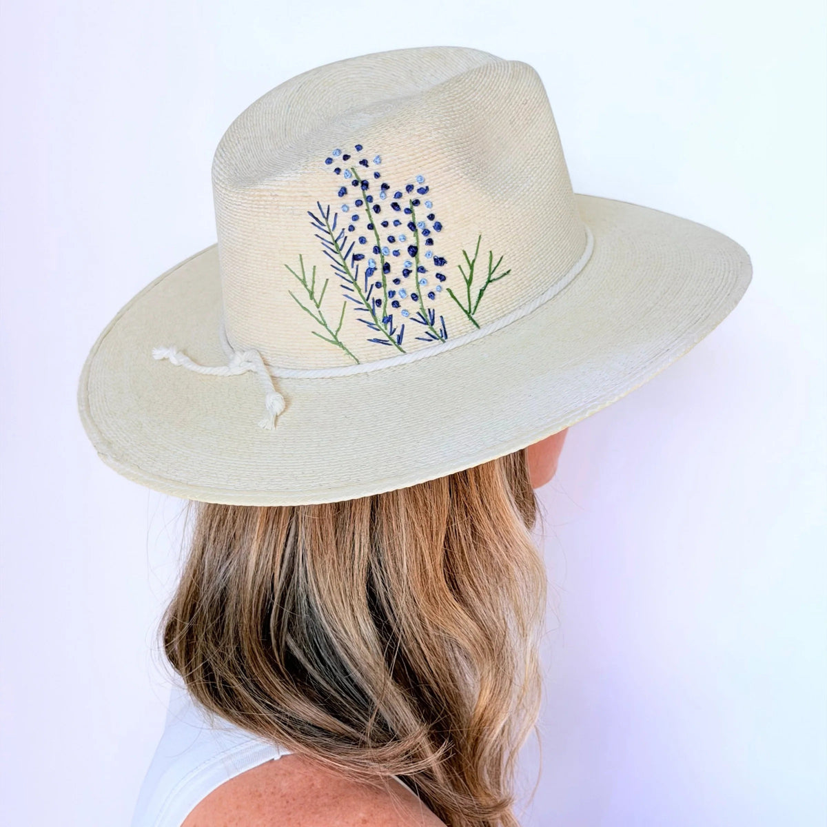 A woman with wavy blonde hair wears a BALDIZ Eggshell Palm Hat, embroidered with blue bonnet flowers, viewed from behind against a plain white background.