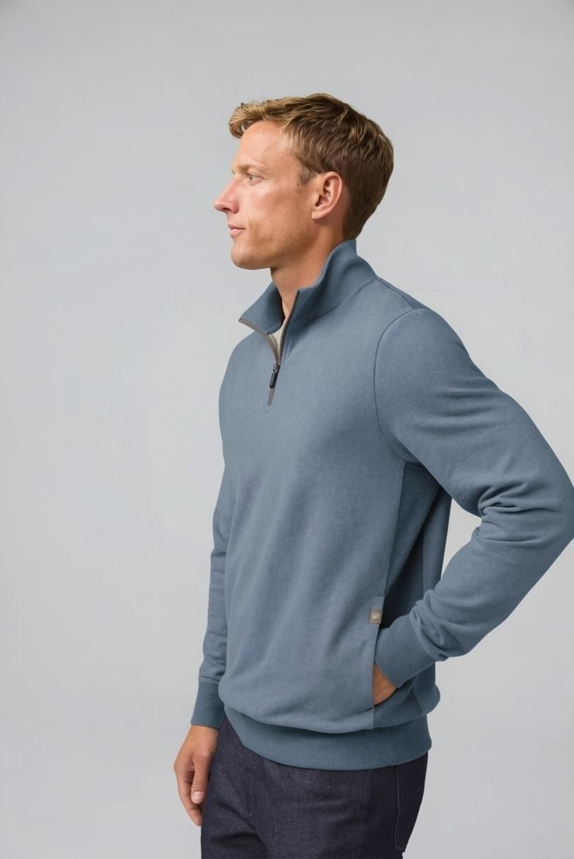 A man with short light brown hair stands in profile, wearing the MACK WELDON Ace 1/2 Zip Sweater in Herringbone and dark pants, one hand in his pocket, against a plain light gray background.