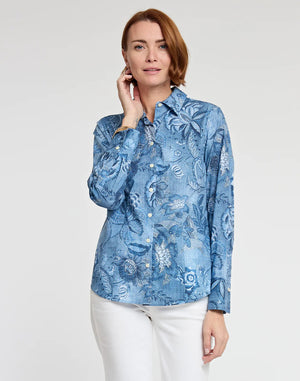 A person with short, reddish-brown hair stands against a plain background, wearing a fitted HINSON WU - DIANE SHIRT IN PASSIONFLOWER PRINT, a blue long-sleeved button-up shirt featuring a light passionflower print pattern, and white pants. The person holds a hand near their face and smiles slightly.