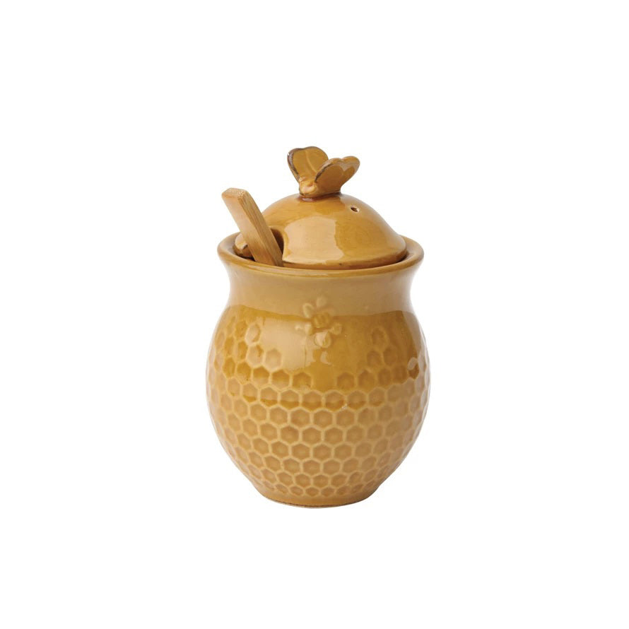 The HONEYCOMB HONEY JAR AND BEE LID by CREATIVE COOP is a ceramic jar featuring an amber-colored, rounded, beehive-like shape with a honeycomb pattern. It comes with a lid adorned with a small bee figurine and includes a wooden honey dipper that rests inside the microwave-safe jar.