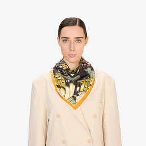 A close-up of a woman wearing a beige blazer and the INOUI EDITIONS - PAPILLON (BUTTERFLIES) Silk Scarf in Green, featuring abstract patterns in yellow, black, green, and white. Only her lower face and neck are visible.