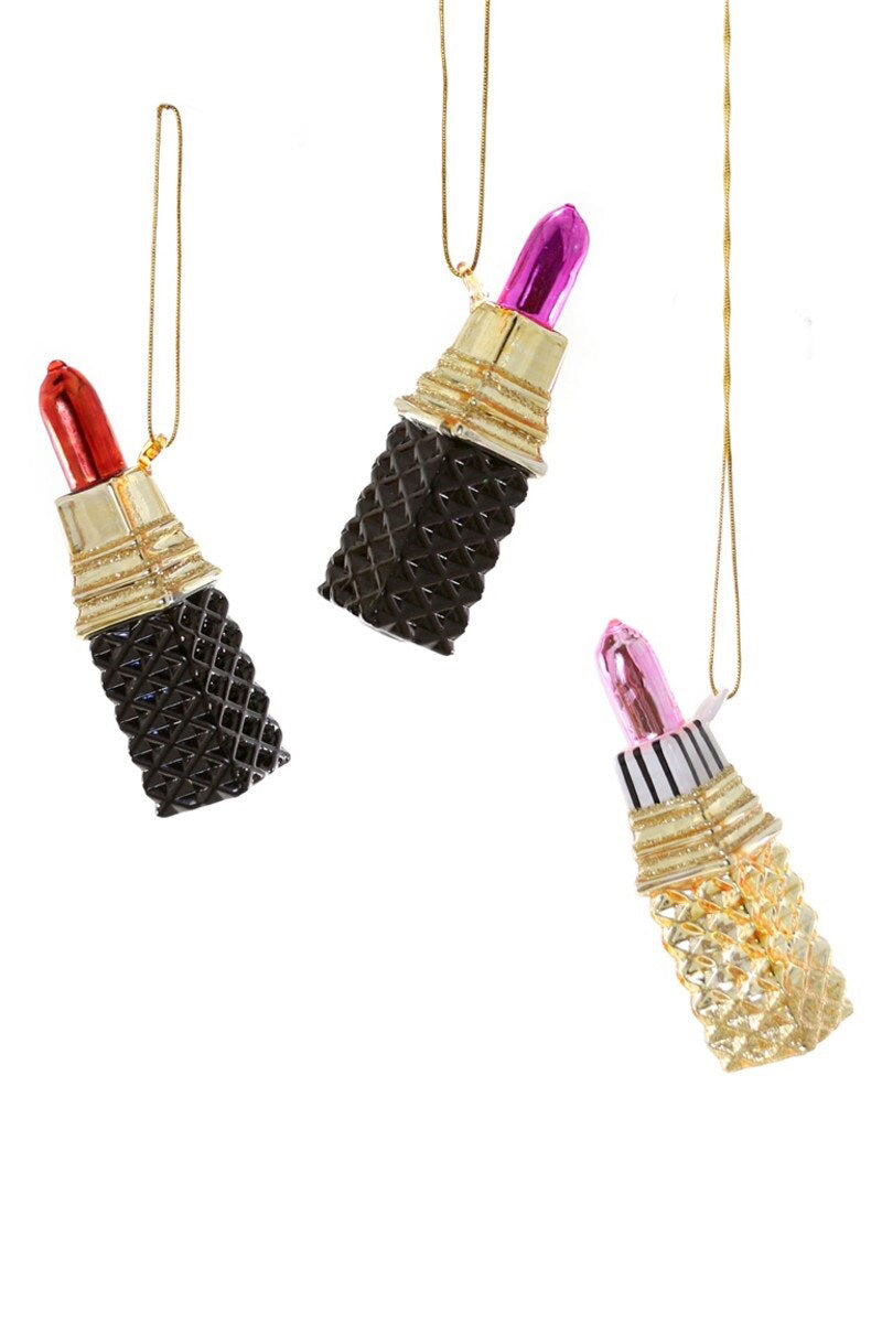The CODY FOSTER LIPSTICK ORNAMENT is a glass ornament shaped like a lipstick, sold individually with a gold string, featuring quilted black or gold bases and metallic red, pink, or purple tops. Call the store for more details.