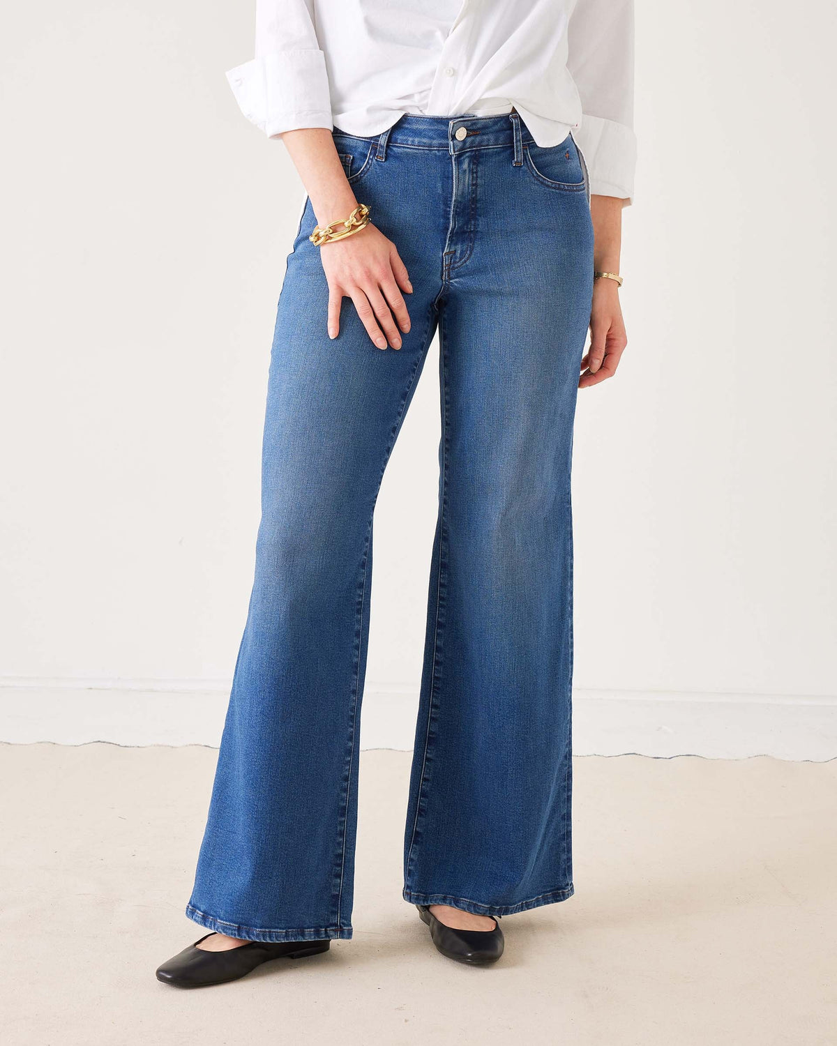 A person wearing the MERSEA Kate Wide Leg Jean in Baltic Wash, featuring four-way-stretch technology, paired with a white long-sleeve shirt with rolled cuffs, black flat shoes, and gold bracelets, stands on a light-colored floor against a plain white background.