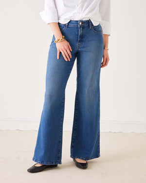 A person wearing the MERSEA Kate Wide Leg Jean in Baltic Wash, featuring four-way-stretch technology, paired with a white long-sleeve shirt with rolled cuffs, black flat shoes, and gold bracelets, stands on a light-colored floor against a plain white background.