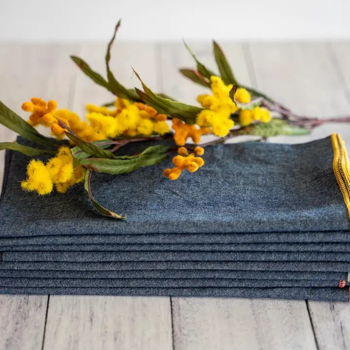 A stack of DOT AND ARMY - CHAMBRAY DINNER NAPKINS rests on a light wooden surface, topped with a sprig of yellow flowers and green leaves.