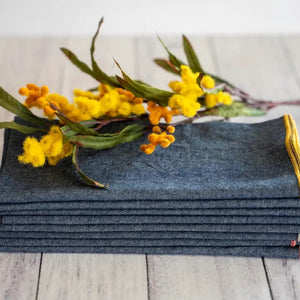 A stack of DOT AND ARMY - CHAMBRAY DINNER NAPKINS rests on a light wooden surface, topped with a sprig of yellow flowers and green leaves.