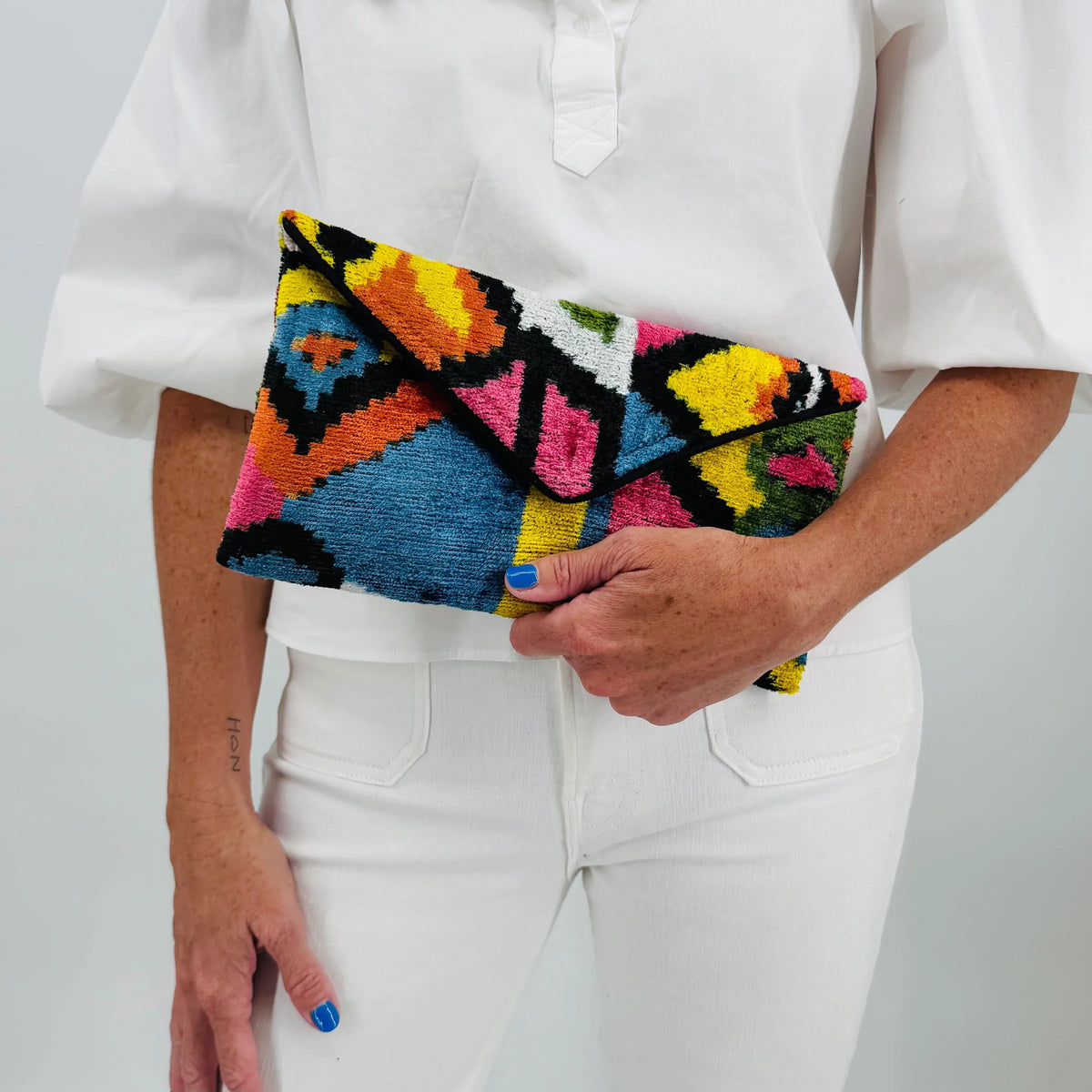 A person in a white outfit holds the BALDIZ - LILY CLUTCH IN PALOMA by BALDIZ, featuring a colorful geometric pattern. Their blue nails and a small forearm tattoo are visible.
