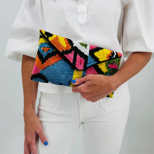 A person in a white outfit holds the BALDIZ - LILY CLUTCH IN PALOMA by BALDIZ, featuring a colorful geometric pattern. Their blue nails and a small forearm tattoo are visible.