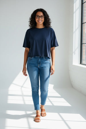 A woman with wavy hair is smiling indoors, standing in front of a white glass-front cabinet and a cozy bench with cushions. She wears the FRANK & EILEEN Perry NYC Crewneck Tee in Vintage Jersey British Royal Navy, styled in a relaxed fit with blue jeans and one hand in her pocket.