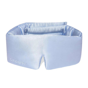 A person wearing a white tank top is lying on a pillow with the DROWSY - SATIN SLEEP MASK by DROWSY SLEEP CO covering their eyes. They look relaxed, enjoying beauty sleep as their hand rests gently on the pillow.