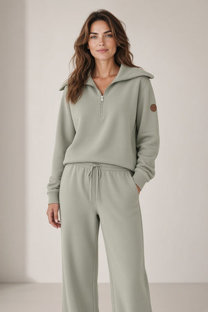 A woman wears a sleek rainy beige SAVE THE DUCK - HINATA HALF ZIP TOP with a wide collar and matching wide-leg drawstring sweatpants.