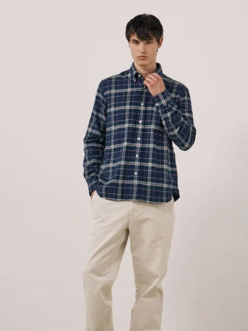 A young person with dark hair, wearing the HARTFORD - PITT CHECKED FLANNEL SHIRT IN BLUE AND GREEN by HARTFORD, looks at the camera while adjusting the shirt collar with one hand against a plain light background.