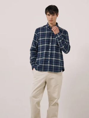 A young person with dark hair, wearing the HARTFORD - PITT CHECKED FLANNEL SHIRT IN BLUE AND GREEN by HARTFORD, looks at the camera while adjusting the shirt collar with one hand against a plain light background.