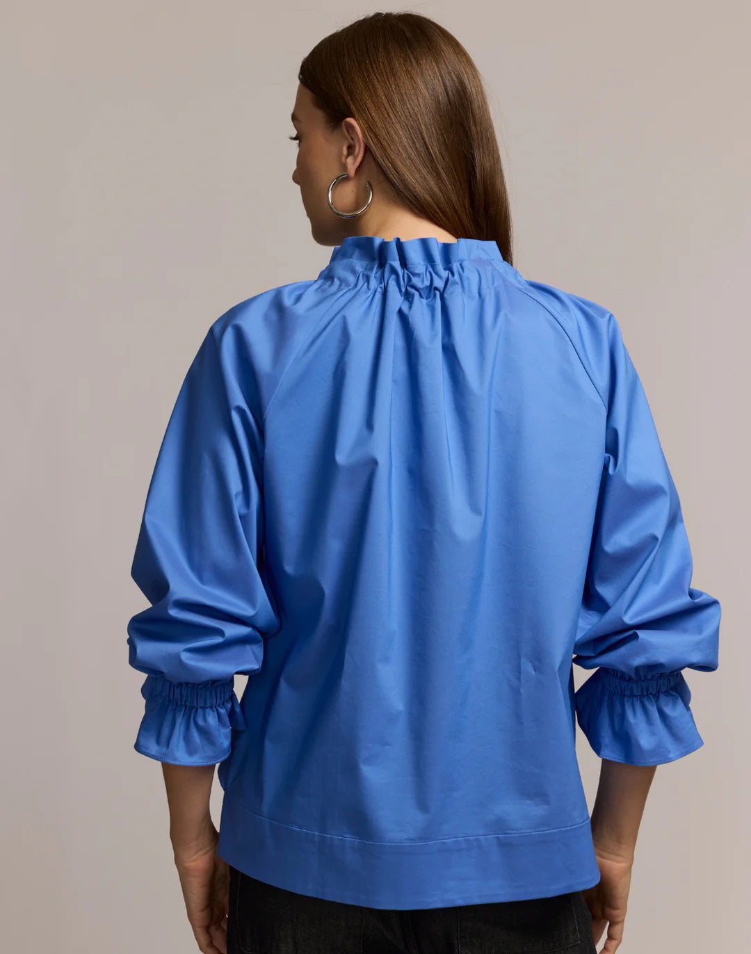 A woman with long brown hair models the HINSON WU ALINA LONG SLEEVE SHIRT, a bright blue blouse with a bow neck and ruffled sleeves, paired with dark pants. She stands against a light background, looking at the camera.