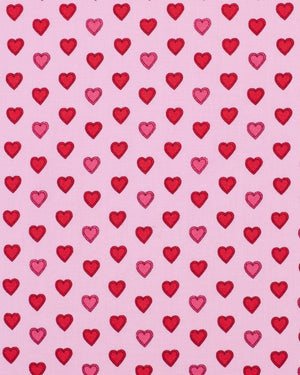 A woman stands barefoot on a wooden floor, wearing the PRINTFRESH - MINI CANDY HEARTS LONG PAJAMA SET by PRINTFRESH. The pajamas feature pink fabric decorated with a red heart pattern. She has long blonde hair and a relaxed expression, posing against a plain white wall.
