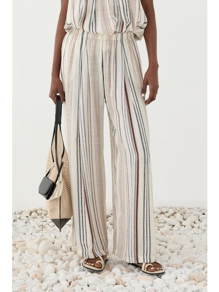 A person stands on white pebbles wearing ELEH’s STRIPED TROUSER—a loose, wide-leg design with a matching sleeveless top in a soft rayon blend—paired with brown sandals and a beige woven bag.