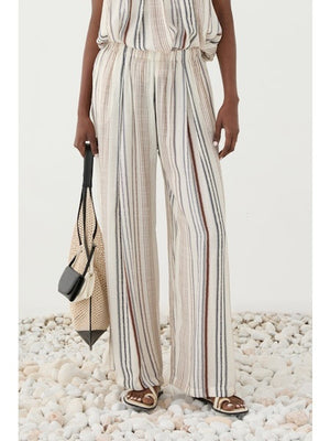 A person stands on white pebbles wearing ELEH’s STRIPED TROUSER—a loose, wide-leg design with a matching sleeveless top in a soft rayon blend—paired with brown sandals and a beige woven bag.