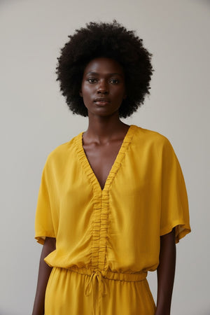 A person with light blonde hair in a low ponytail stands with their back to the camera, wearing the NUDE Tie Waist V Neck Blouse by NUDE in yellow, set against a plain brown background.