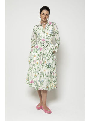 Against a plain background, someone models the LAVI COUTURE SPRITZ DRESS by LAVI, featuring green and pink botanical prints. This long Italian garden dress has long sleeves, a tied belt, and pockets, all crafted from breathable cotton. Their pink shoes complete the look elegantly.