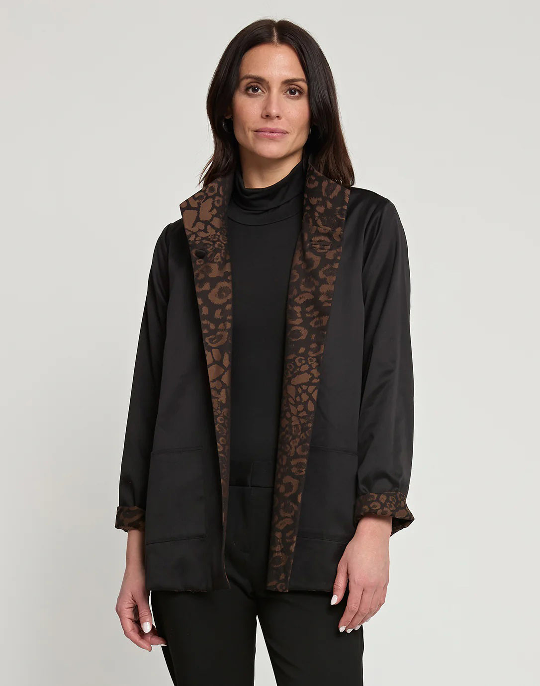 A woman with long dark hair is wearing the HINSON WU Constance Reversible Jacket, which features stylish leopard print details on the collar and belt. She pairs it with a black turtleneck top and pants while standing against a light background. The look balances style with practicality, as the jacket is conveniently machine washable.