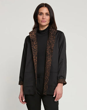 A woman with long dark hair is wearing the HINSON WU Constance Reversible Jacket, which features stylish leopard print details on the collar and belt. She pairs it with a black turtleneck top and pants while standing against a light background. The look balances style with practicality, as the jacket is conveniently machine washable.