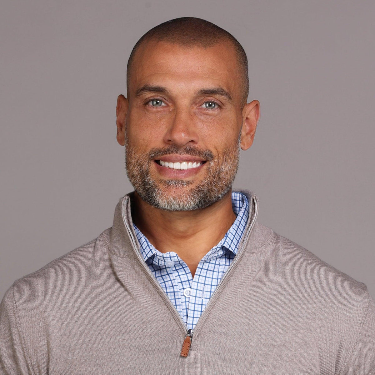 A smiling man with a shaved head and trimmed beard wears the COLLARS AND CO Oatmeal Cashmere Touch Australian Merino Quarter Zip Sweater over a blue checkered shirt, posing against a plain gray background.