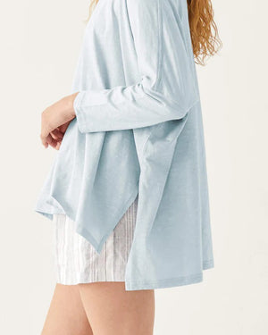 A woman with wavy brown hair models the MERSEA - CATALINA SLUB TEE—an oversized, long-sleeved white top with thin gray stripes—by MERSEA. She pairs it with blue jeans and stands against a plain, light background, hands behind her back.