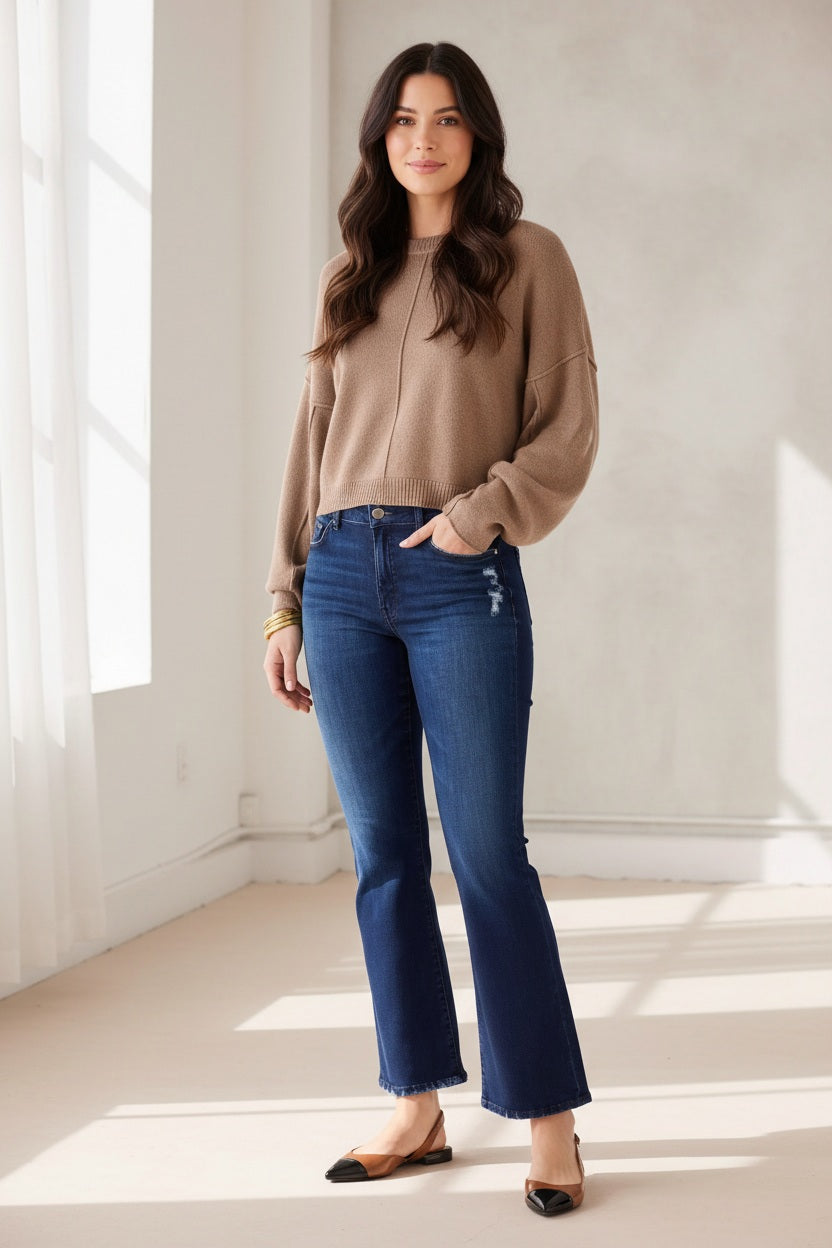 A person stands with hands in pockets against a plain light background, wearing MERSEA’s Murphy Mini Boot Jeans in Patriot—a pair of mid-rise, blue flare jeans crafted from four-way stretch denim—paired with a tan long-sleeve top, black pointed-toe flats, and gold bangles.