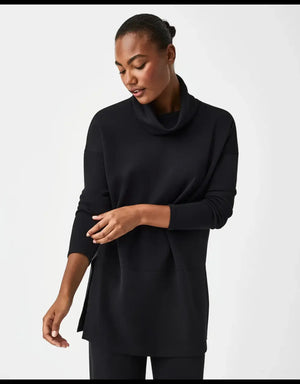A person pulls up the collar of the SPANX - AIRESSENTIALS TURTLENECK TUNIC IN BLACK, smiling with closed eyes against a plain light background—perfectly showcasing the brand's light-as-air fabric and playful style.