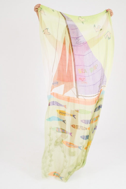 A woman with shoulder-length blonde hair wears a light pink shirt and the STORIATIPIC - GERTRUD SCARF IN ANISE, a colorful modal and silk accessory featuring green, yellow, and purple tones. She stands against a plain light background, looking directly at the camera.
