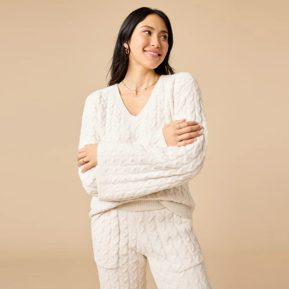 A woman wearing the SOFTIES CABLE KNIT MARSHMALLOW V-NECK WITH FLARED SLEEVE AND PANT SET, a marshmallow-soft, cream-colored cable knit v-neck sweater with wide sleeves and matching lounge pants.