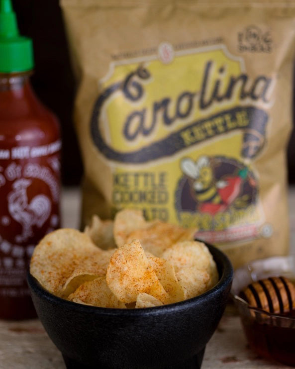 A 5-ounce bag of CAROLINA KETTLE - HONEY SRIRACHA CHIPS from 1 IN 6 SNACKS. The packaging features a vibrant design with a bee mascot holding a dripping honey stick and highlights the sweet and hot flavor, as well as emphasizing the natural crispiness and guaranteed freshness.
