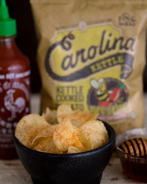 A 5-ounce bag of CAROLINA KETTLE - HONEY SRIRACHA CHIPS from 1 IN 6 SNACKS. The packaging features a vibrant design with a bee mascot holding a dripping honey stick and highlights the sweet and hot flavor, as well as emphasizing the natural crispiness and guaranteed freshness.