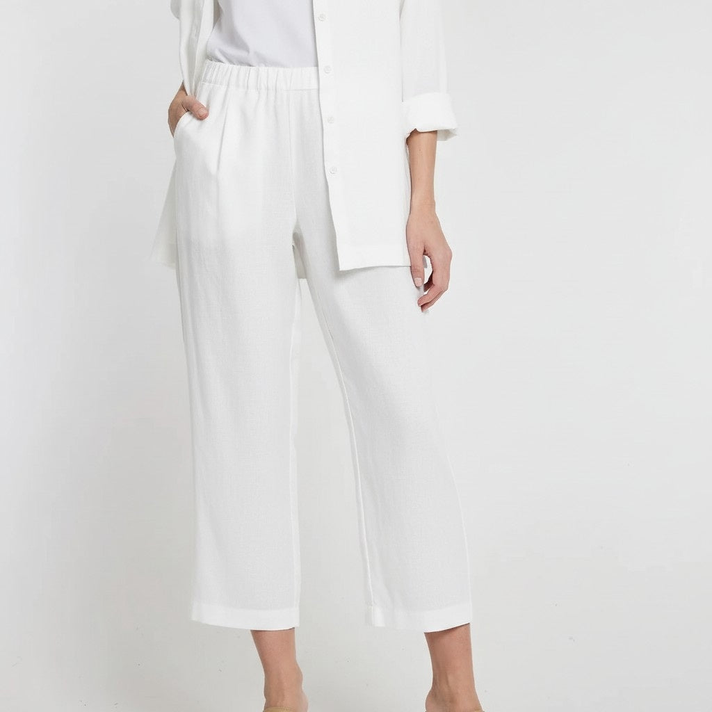 A person wearing HINSON WU - BIANCA LUXE LINEN PANT, a loose-fitting, cropped white 100% linen pant with an elastic waistband from HINSON WU, along with a white shirt and top, stands against a plain white background with hands in their pockets.