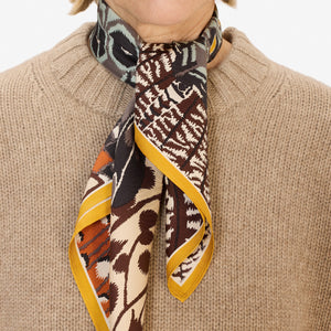 A close-up of a person wearing the INOUI EDITIONS - PAPILLON (BUTTERFLIES) SQUARE WOOL SCARF IN NATURAL by INOUI, draped around their neck over a black top. The scarf showcases abstract and geometric butterfly designs in earthy tones.