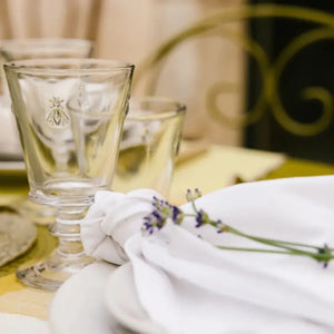 The LA ROCHERE BEE WATER GLASS, with its embossed Napoleonic bee, rests on a sunlit table beside a green lime drink, fresh cantaloupe slices, and white flowers for an effortlessly elegant display.