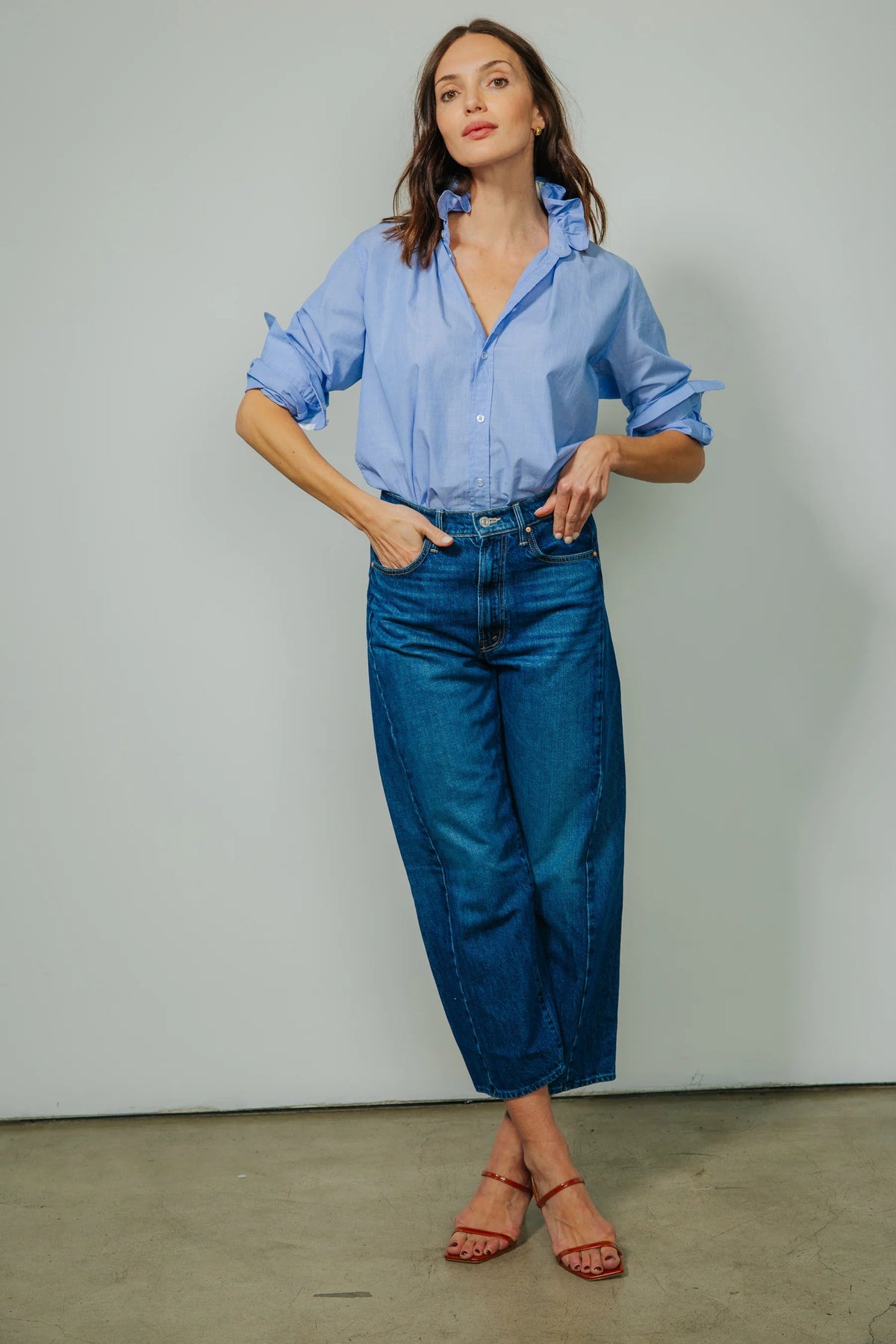 A woman with wavy brown hair wears the KMJ - Quarantina Shirt in Chambray, its feminine ruffle collar and relaxed fit highlighted as she stands with one hand in her pocket against a plain light gray background, looking confidently at the camera.
