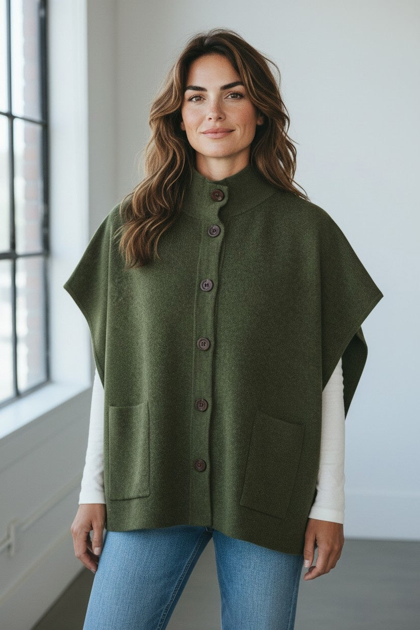WOMAN wearing the KINROSS CASHMERE Short Sleeve Button Mock Cardigan in Highland layered over a white long-sleeve top and jeans, standing indoors near a large window and smiling softly at the camera. Her look is completed with this oversized sleeveless cardigan by KINROSS for added warmth and style.