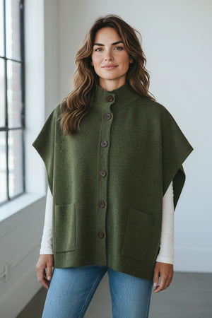 WOMAN wearing the KINROSS CASHMERE Short Sleeve Button Mock Cardigan in Highland layered over a white long-sleeve top and jeans, standing indoors near a large window and smiling softly at the camera. Her look is completed with this oversized sleeveless cardigan by KINROSS for added warmth and style.