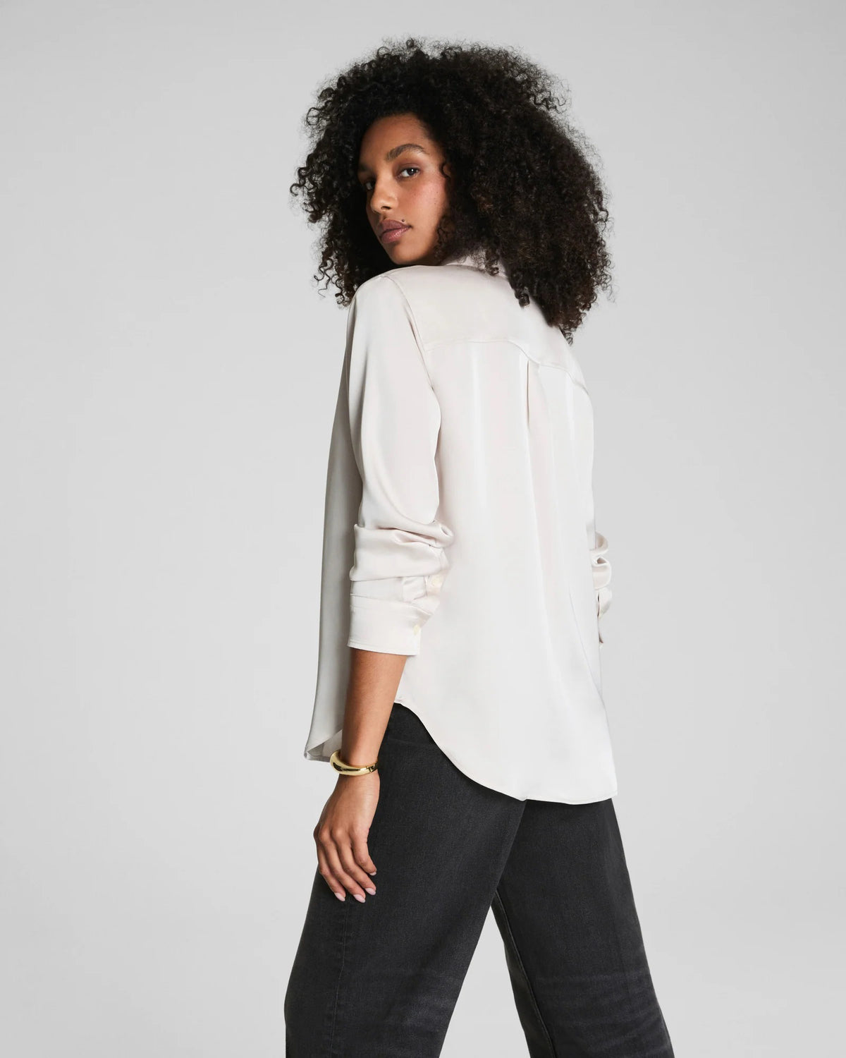 A woman with curly hair wears the SPANX Silky Smooth Button Down Shirt in a relaxed tailored fit and black pants, turning slightly as she looks over her shoulder against a plain gray background.