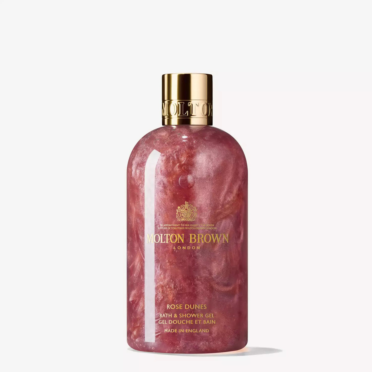 The MOLTON BROWN - ROSE DUNES SHOWER GEL features a transparent bottle showcasing a rose-colored, shimmering gel. It is adorned with a gold cap and a gold label bearing the Molton Brown London name. This sleek, ambery body wash stands out against a white background.