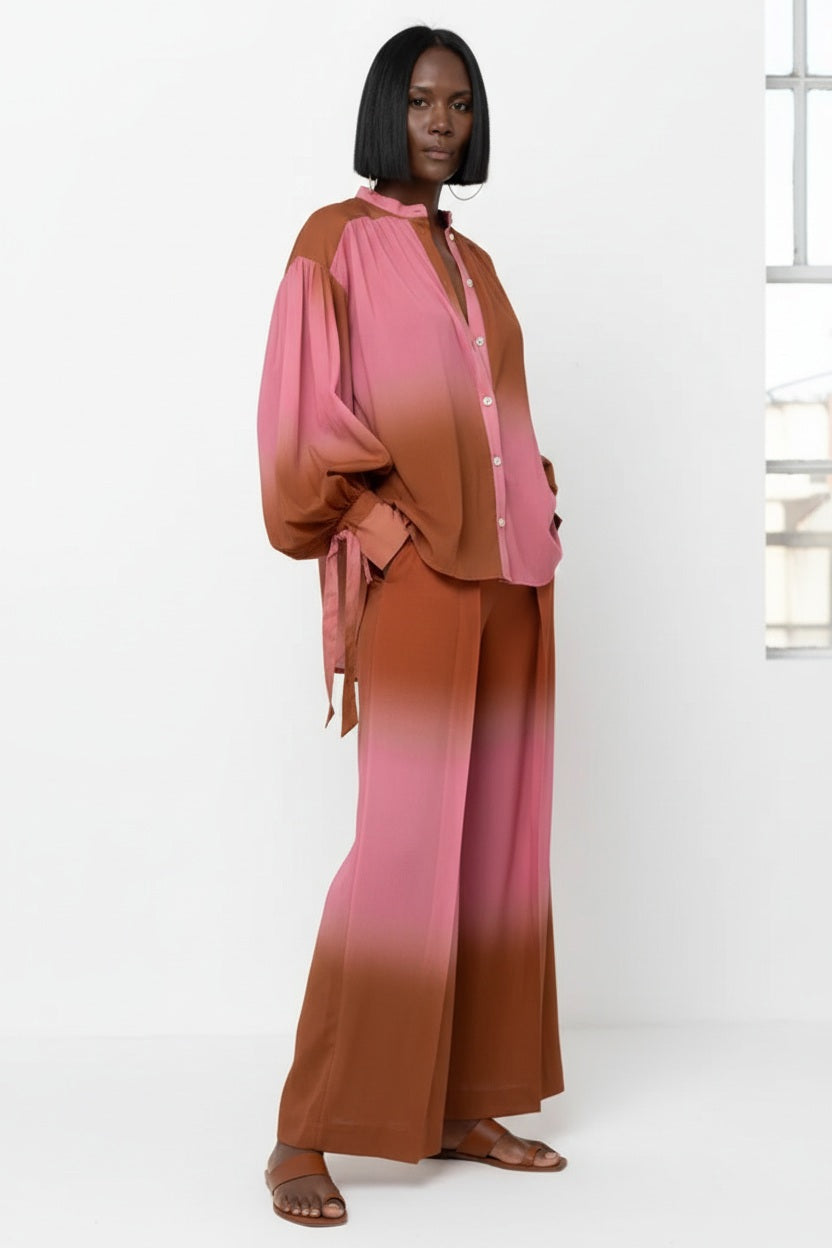 A person with straight black hair models the HOPE FOR FLOWERS WRIST TIE BLOUSE by HOPE FOR FLOWERS, featuring a flowy design in a pink and brown ombre, highlighted as sustainable clothing against a plain white background.