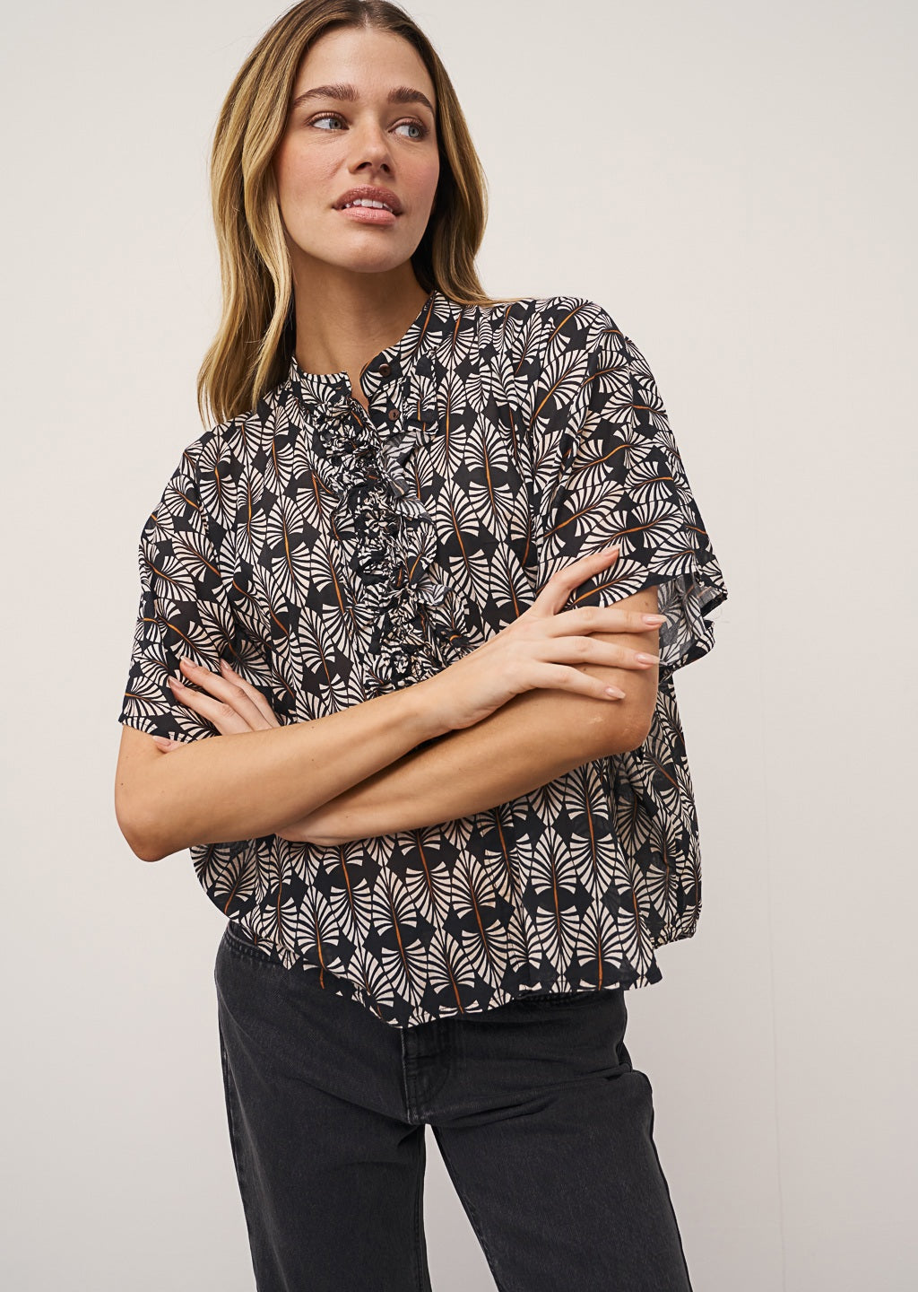 A woman stands against a light wall, wearing the DAISY BLOUSE IN BLACK AND WHITE PRINT by 0039 ITALY, black wide-leg jeans with frayed hems, and black mesh flats. One hand is in her pocket and she looks relaxed.
