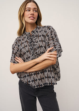 A woman stands against a light wall, wearing the DAISY BLOUSE IN BLACK AND WHITE PRINT by 0039 ITALY, black wide-leg jeans with frayed hems, and black mesh flats. One hand is in her pocket and she looks relaxed.