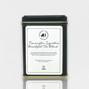 The Fearrington Signature Collection's black tin container features a white label with the text "Fearrington Signature Breakfast Tea Blend" from Fearrington House. The label describes this loose leaf tea as a rich, robust blend of Ceylon and Assam. The bottom of the tin bears a logo and text indicating that it is handmade.