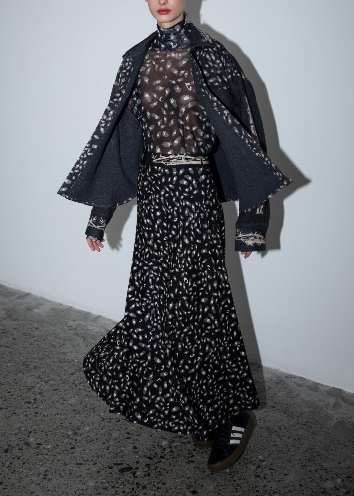 A person stands on a concrete floor wearing the FRANCO FERRARI - JEMMA WOOL MIDI SKIRT, a dark patterned jacket over their shoulders, a sheer patterned top, and black-and-white shoes. Their face is partially out of frame.