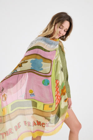 A smiling woman with wavy blonde hair wears a light green textured top and the STORIATIPIC GABIE SCARF IN ROSE, a colorful cotton-modal accessory with abstract patterns, against a plain white background.