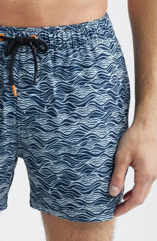 A shirtless man stands with hands in his pockets against a plain light background, modeling SAVE THE DUCK's Ademir Swim Trunks in Ondine Off White on Blue, crafted from quick-dry recycled polyester.