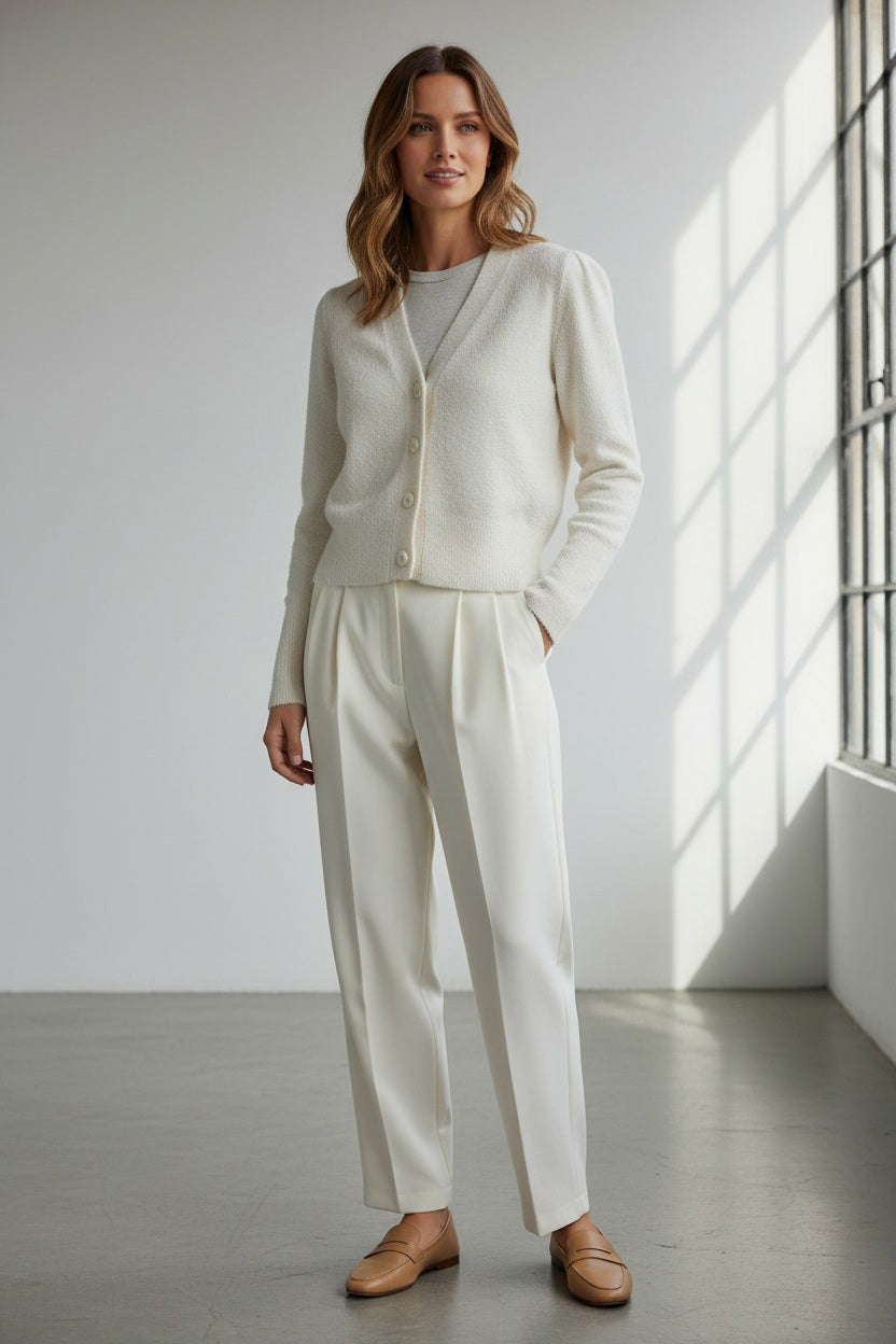 A woman with straight black hair wears the KINROSS CASHMERE Stretch Boucle Button Cardigan by KINROSS and white pants, standing against a plain white background with one hand in her pocket, facing the camera and smiling softly.
