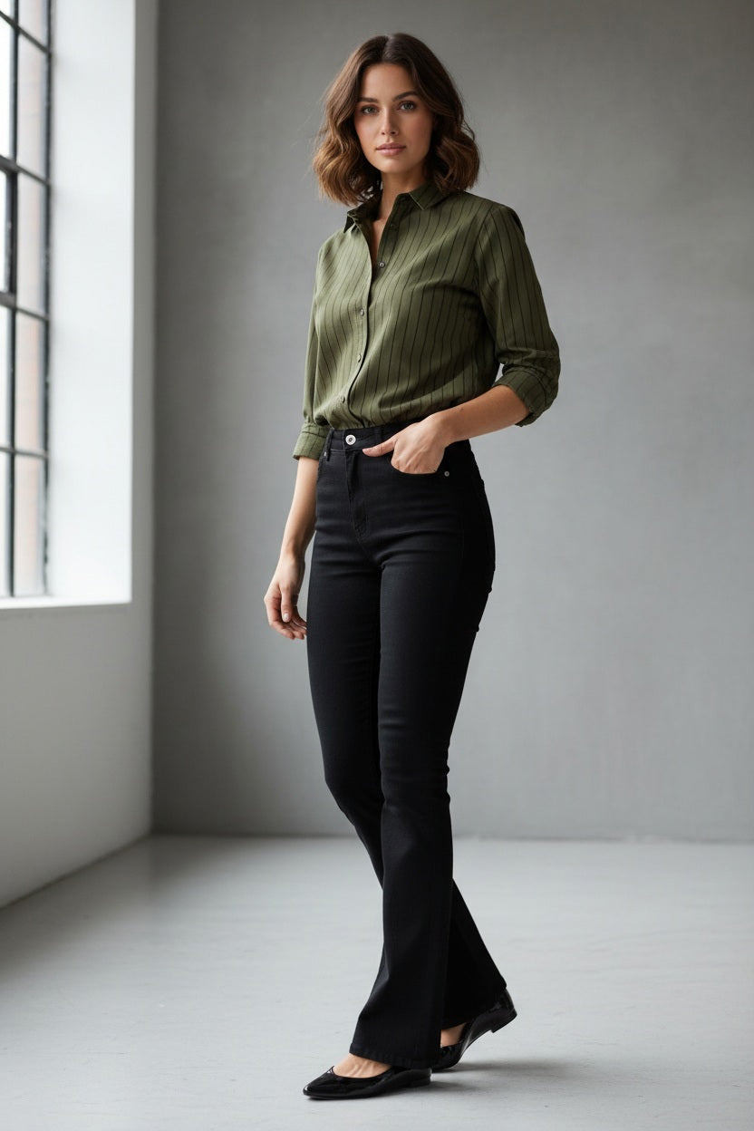 A woman stands indoors near a window, wearing a green button-up shirt styled with SPANX Pull On Flare Jean in Obsidian Rinse and black shoes. The background is minimal and gray.